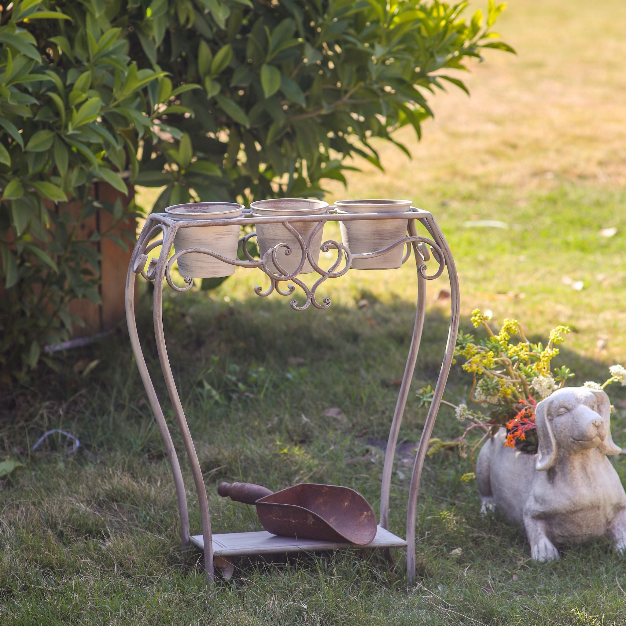 Yolande three pot plant stand in distressed taupe metal with scrolled frame and lower shelf, empty pots, in a garden. 